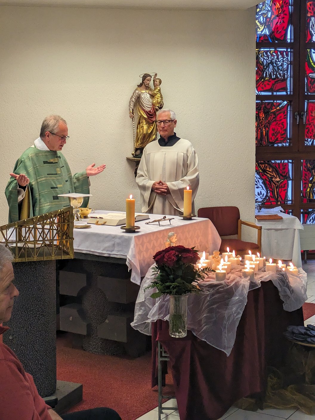 Zwei Geistliche in liturgischer Kleidung stehen an einem Altar mit Kerzen und Blumen in einer Kirche mit buntem Kirchenfenster.