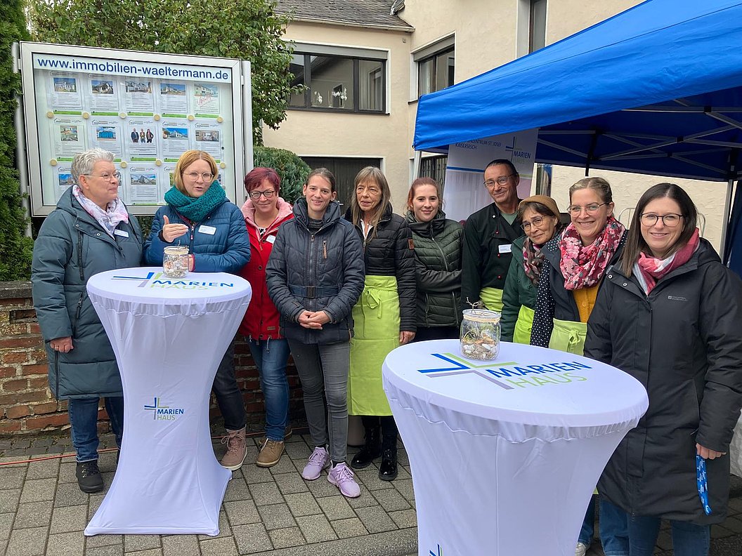 Gruppe von Menschen steht hinter zwei Stehtischen mit weißen Tischdecken und dem Schriftzug "Marien" im Freien vor einem Immobilienbüro.