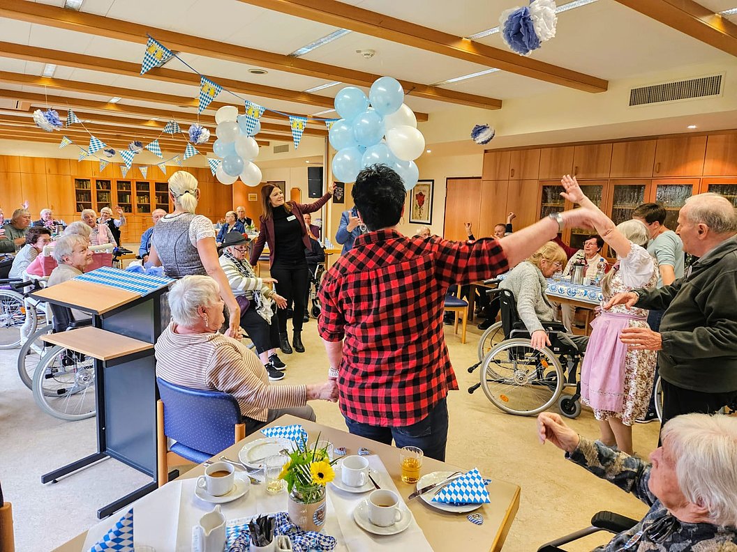 Seniorinnen und Senioren in einem festlich geschmückten Raum mit blau-weißen Luftballons und Girlanden, einige sitzen im Rollstuhl, andere stehen und halten sich an den Händen oder klatschen